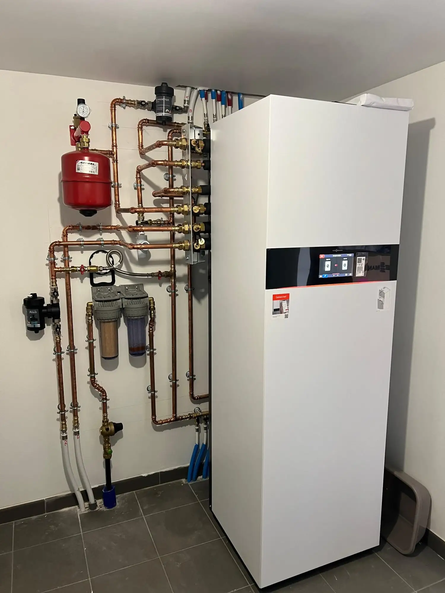 Module intérieur blanc de pompe à chaleur dans un local technique carrelé, réseau en cuivre, vase d’expansion rouge et filtres sur les tuyaux.