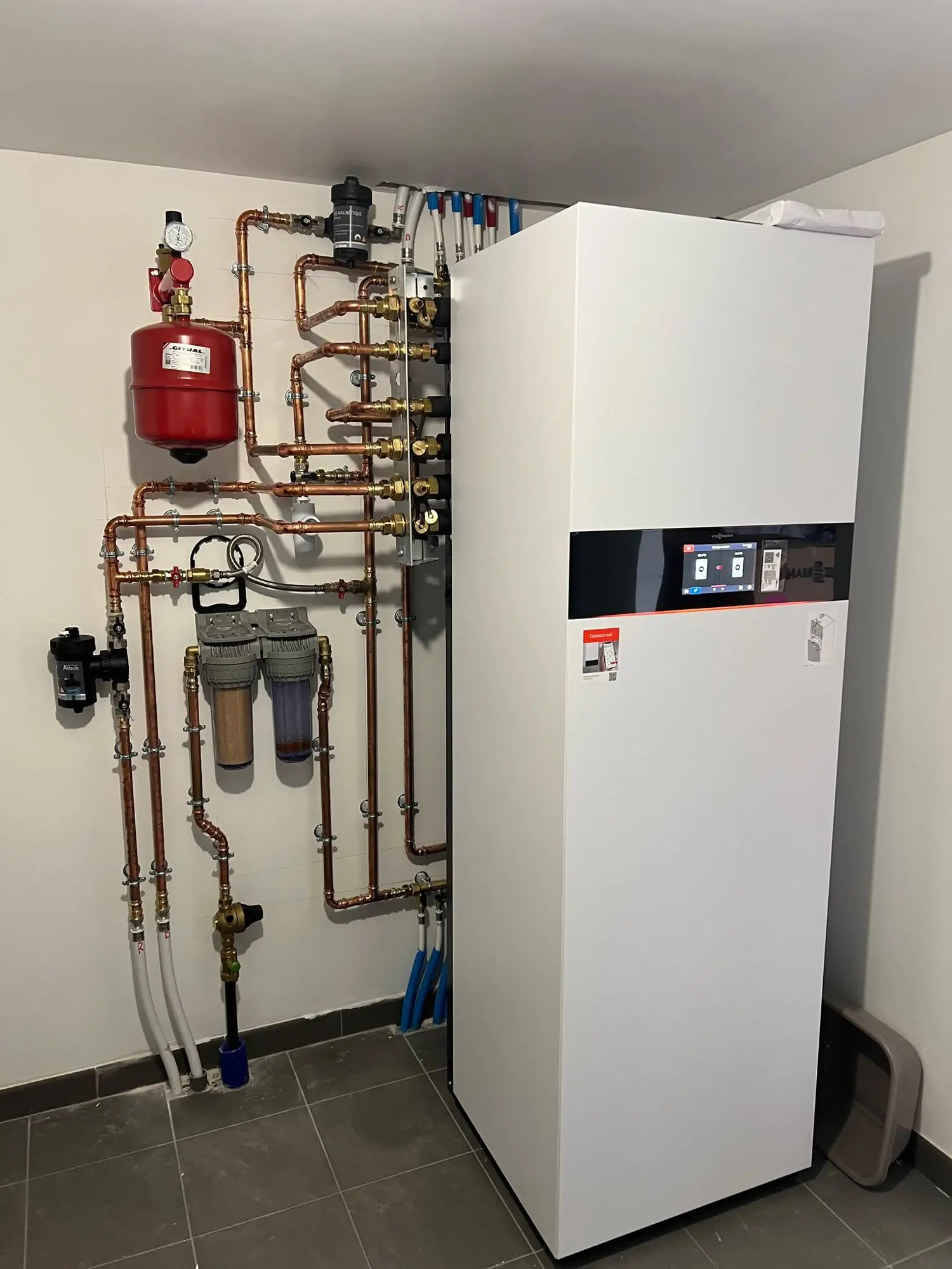Module intérieur blanc de pompe à chaleur dans un local technique carrelé, réseau en cuivre, vase d’expansion rouge et filtres sur les tuyaux.