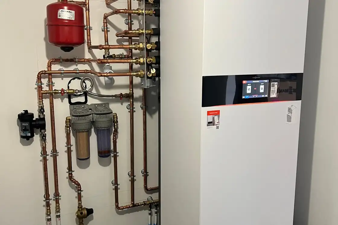 Module intérieur de pompe à chaleur raccordé à un réseau en cuivre, avec vase d’expansion rouge et filtres, dans un local technique carrelé.