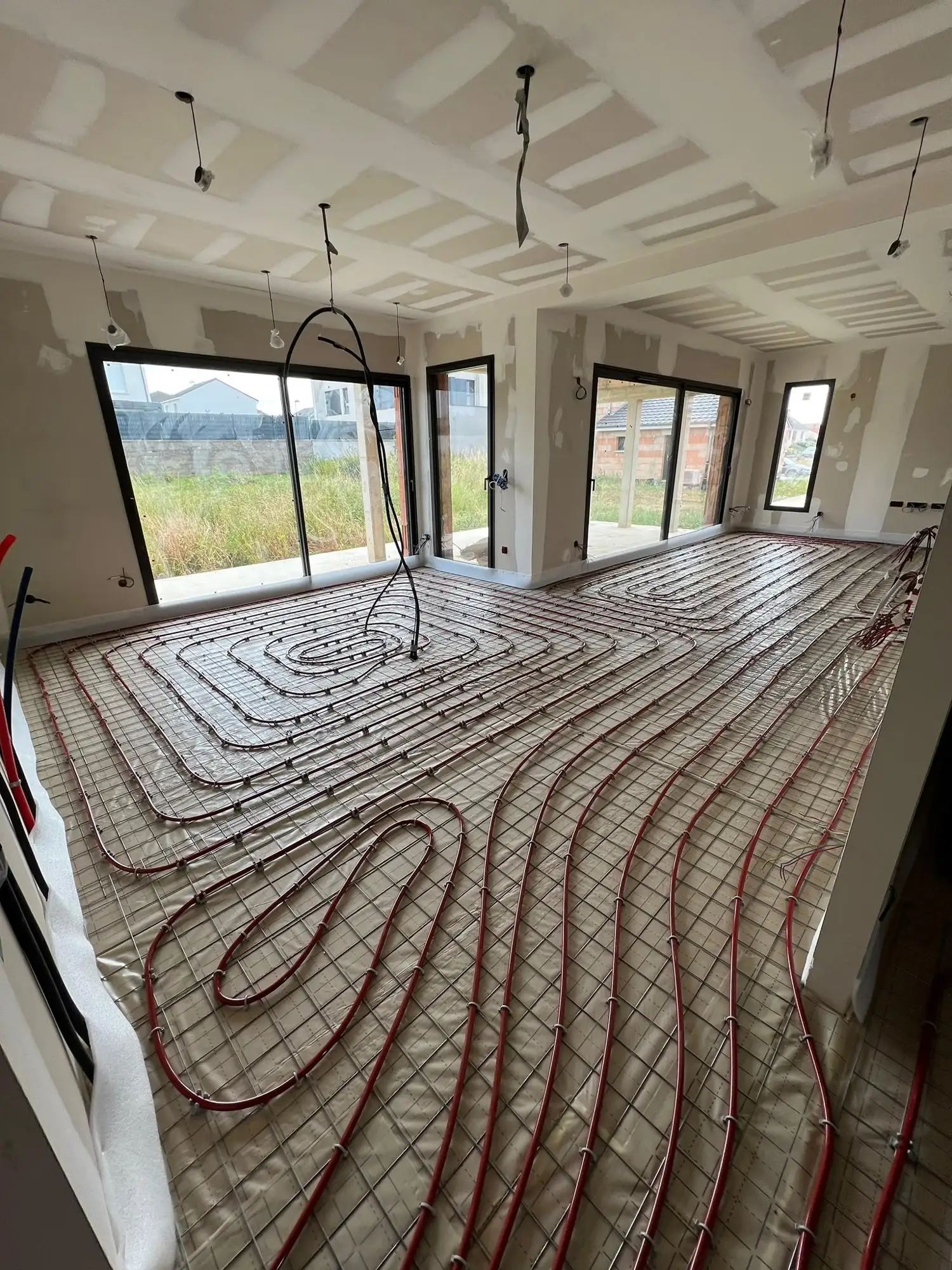 Grande pièce de maison neuve aux baies vitrées, tubes rouges de plancher chauffant posés en spirale sur un isolant quadrillé, bande périphérique blanche le long des murs.