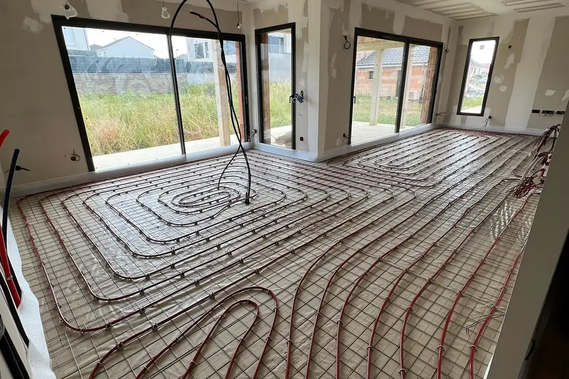 Tubes de plancher chauffant posés en spirale sur un isolant quadrillé dans une grande pièce de maison neuve aux baies vitrées.