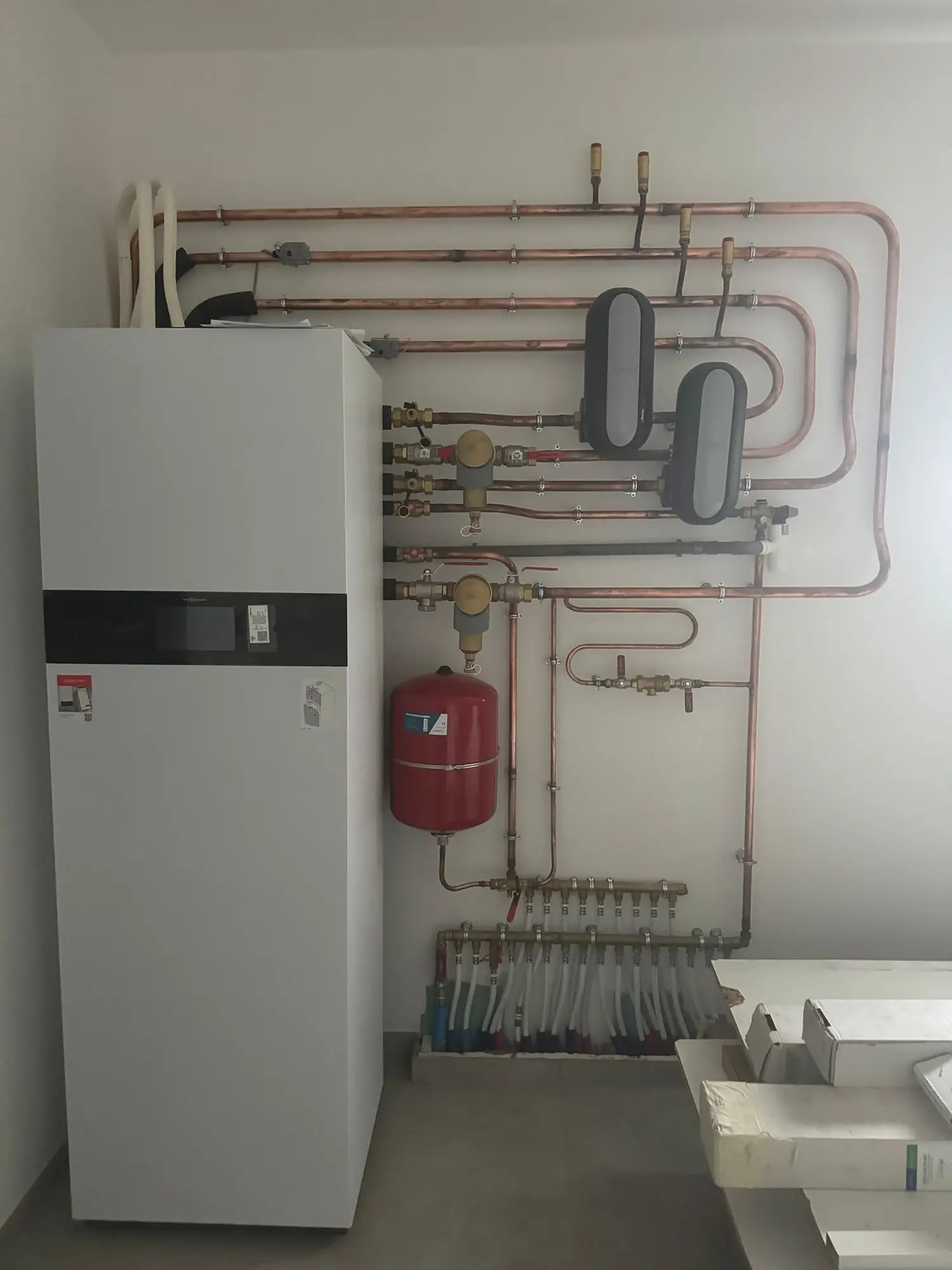 Module intérieur de pompe à chaleur dans un local technique, réseau en cuivre fixé au mur, vases d’expansion et collecteurs de plancher chauffant.