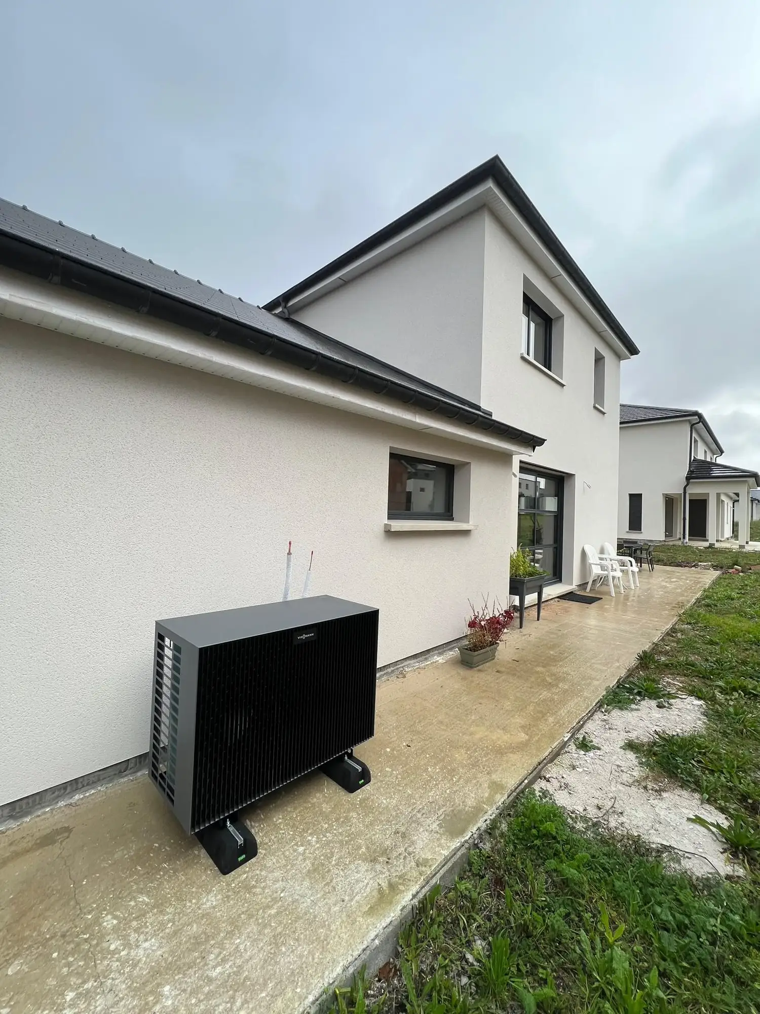 Maison récente à façade enduite blanche, unité extérieure noire de pompe à chaleur posée sur plots au pied du mur, pelouse.