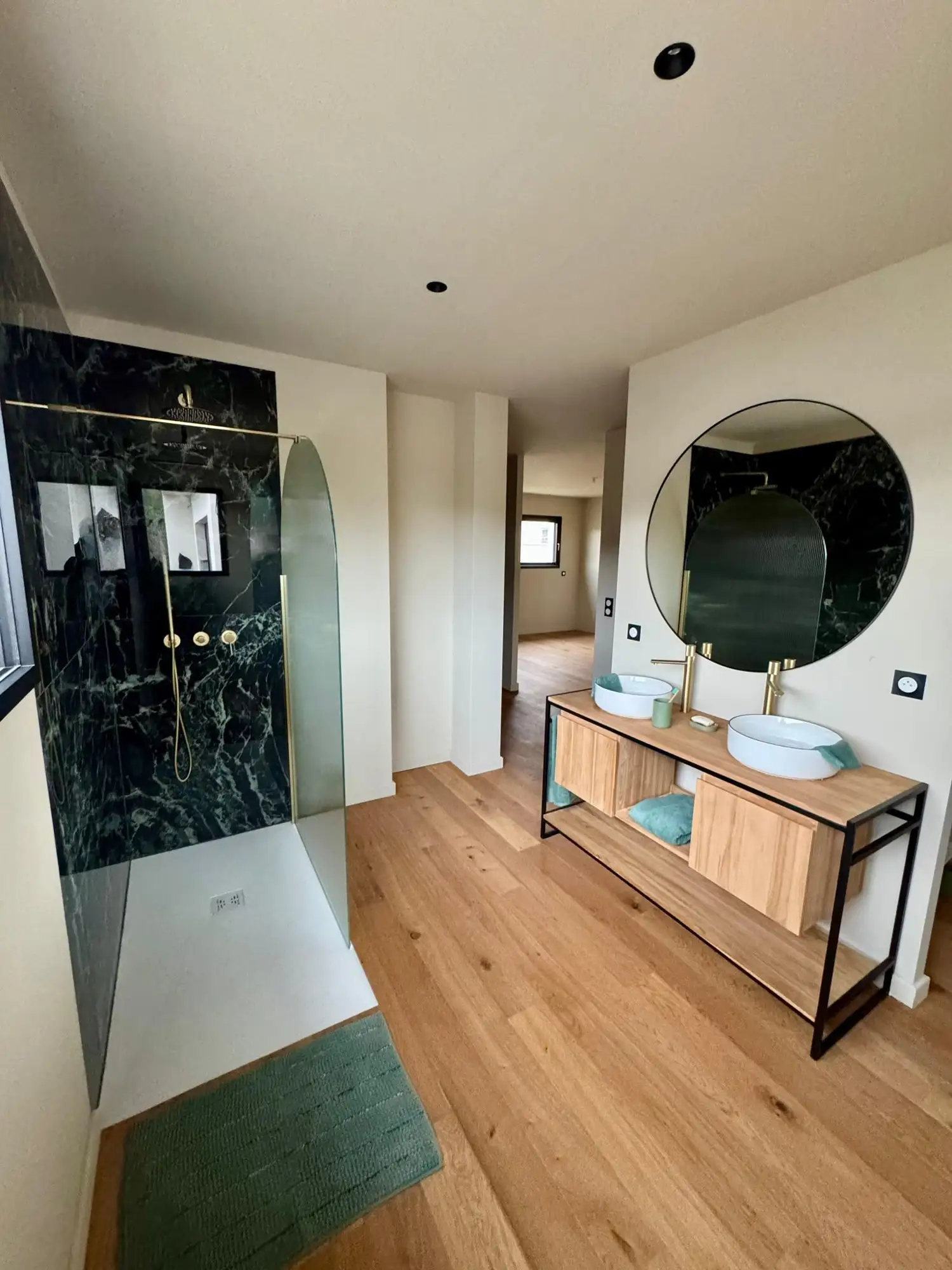 Salle de bains au parquet clair, double vasque à poser sur un meuble en bois, miroir rond, douche à paroi vitrée arrondie devant un mur vert foncé.