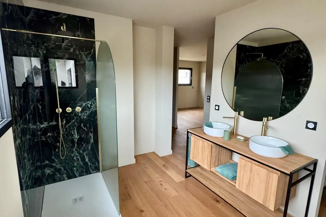 Salle de bains rénovée avec double vasque sur meuble bois, miroir rond, douche à paroi vitrée et mur vert foncé.