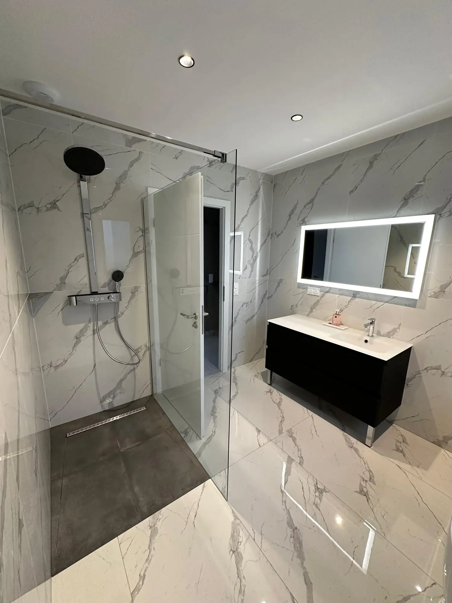 Douche à l’italienne avec paroi vitrée fixe et caniveau de sol, colonne de douche, carrelage effet marbre, meuble vasque noir et miroir lumineux.
