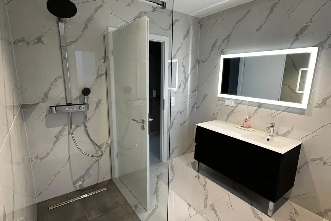 Douche à l’italienne avec paroi vitrée, carrelage effet marbre, meuble vasque noir et miroir lumineux.