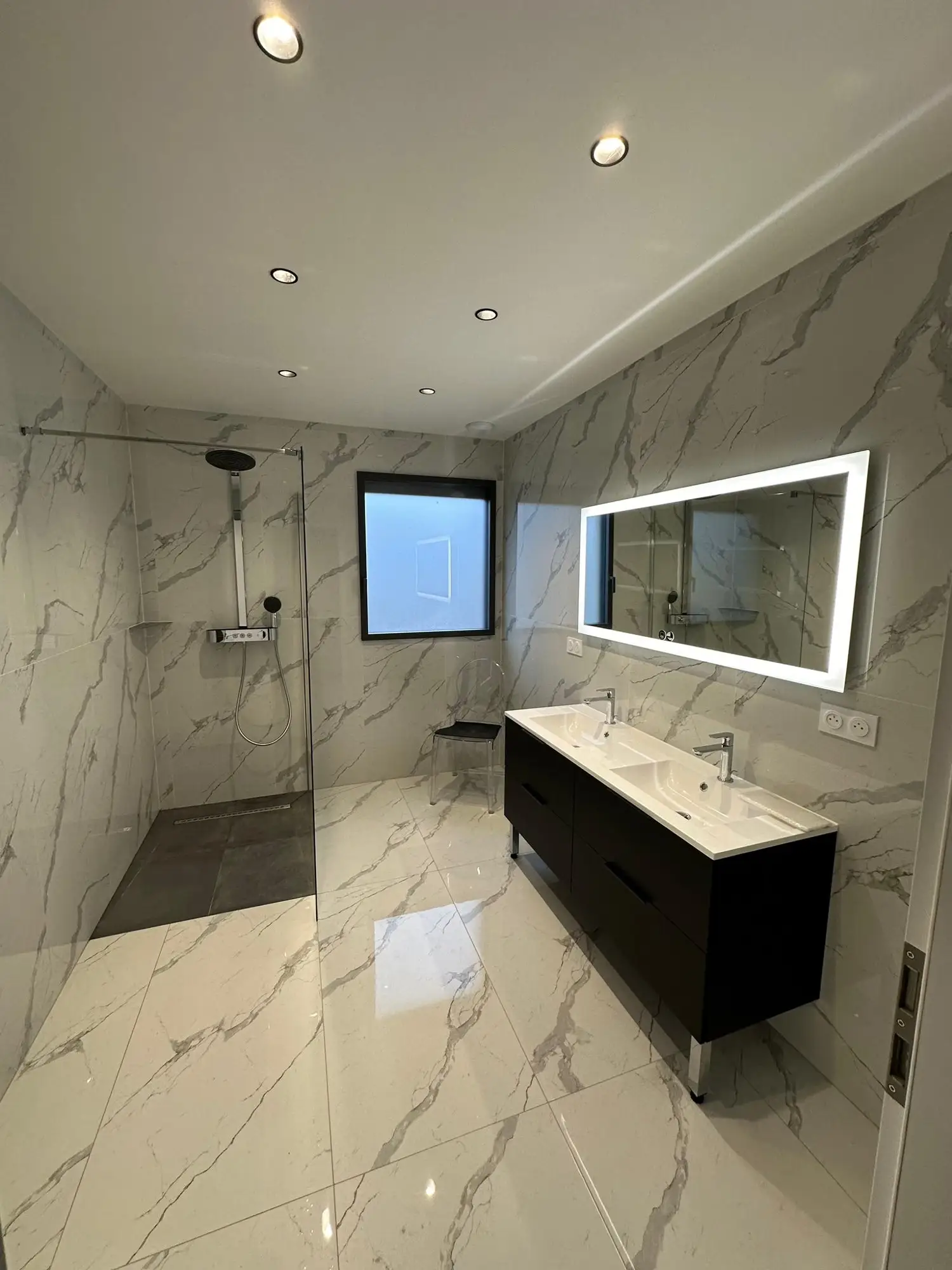 Salle de bains aux grands carreaux effet marbre, douche à l’italienne à sol anthracite, meuble double vasque noir suspendu et miroir rétroéclairé.